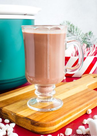 A tall, stemmed glass mug of creamy hot chocolate, garnished with a dusting of cocoa and standing on a wooden cutting board with a red background. Small white marshmallows and a candy cane are visible in the foreground.