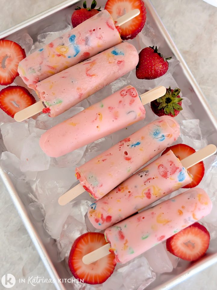 Overhead view of four pink yogurt popsicles studded with rainbow cereal, arranged diagonally on crushed ice in a metal tray and garnished with sliced fresh strawberries.