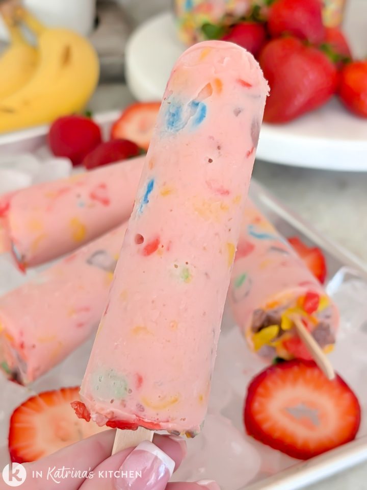 Close-up shot of a hand holding a pink yogurt and cereal popsicle on a wooden stick, with fresh strawberry slices and additional popsicles resting on crushed ice in the background.