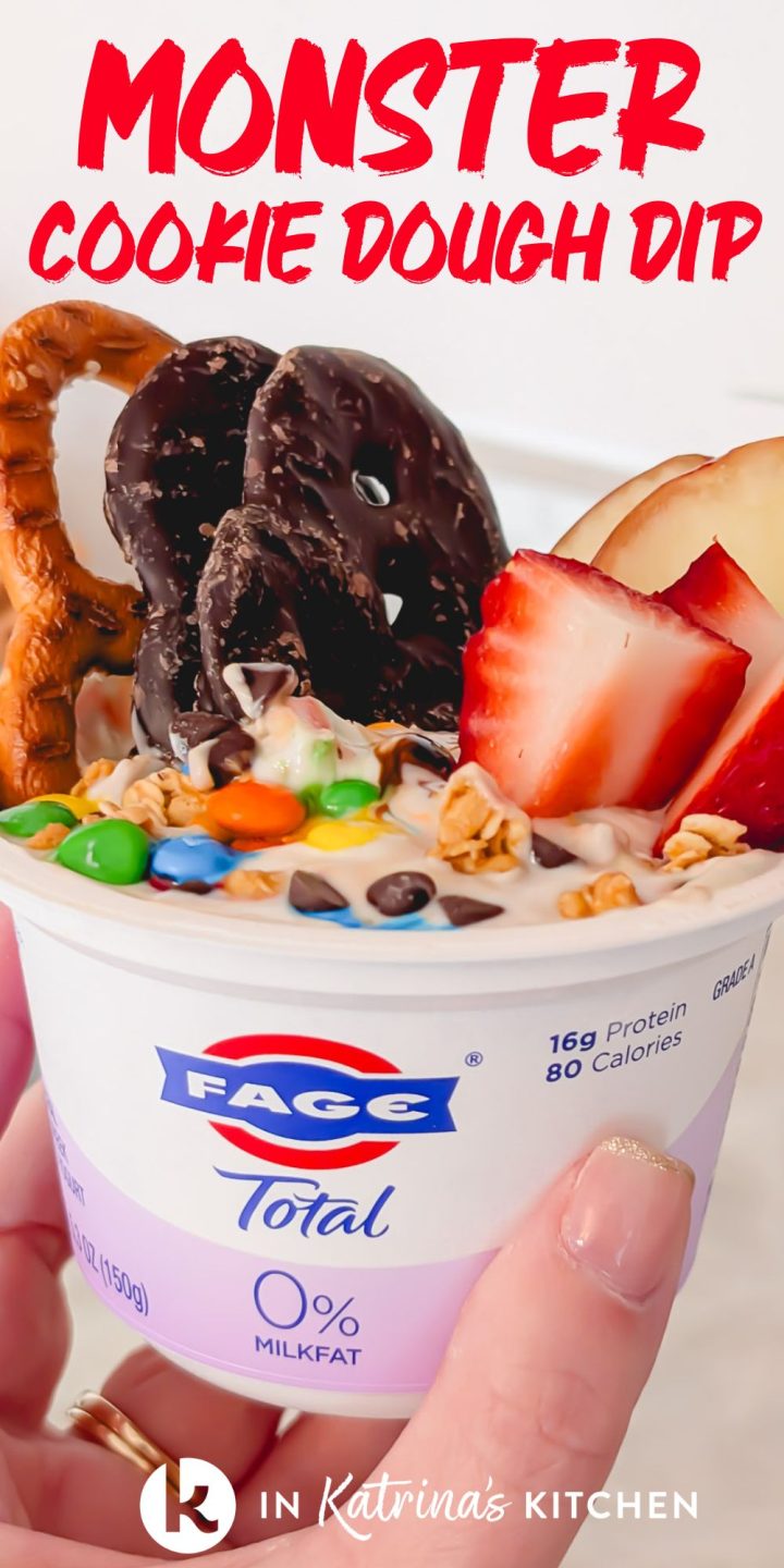 A hand holding a single-serve container of FAGE Total 0% Greek yogurt filled with Monster Cookie Dough dip, topped with mini M&Ms, chocolate chips, pretzel twists, sliced strawberries, and a chocolate cookie, with the text "MONSTER COOKIE DOUGH DIP" overlaying the top of the image.