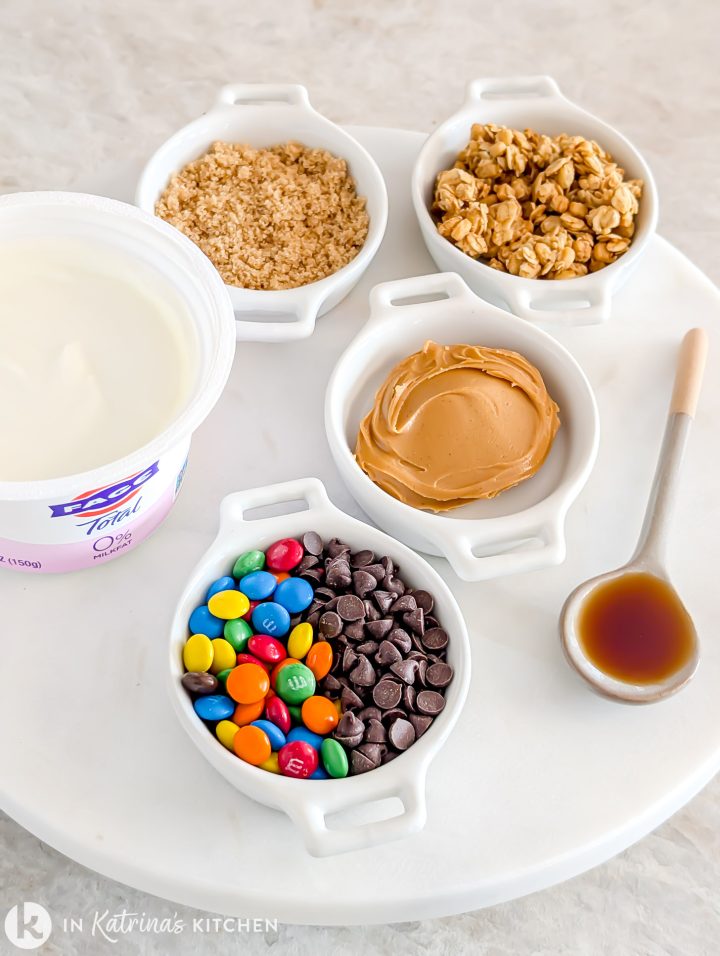 Top-down view of ingredients for Monster Cookie Dough Dip arranged on a white tray, including FAGE 0% Greek yogurt, creamy peanut butter, granola, mini chocolate chips, mini M&Ms, and a spoon of vanilla.