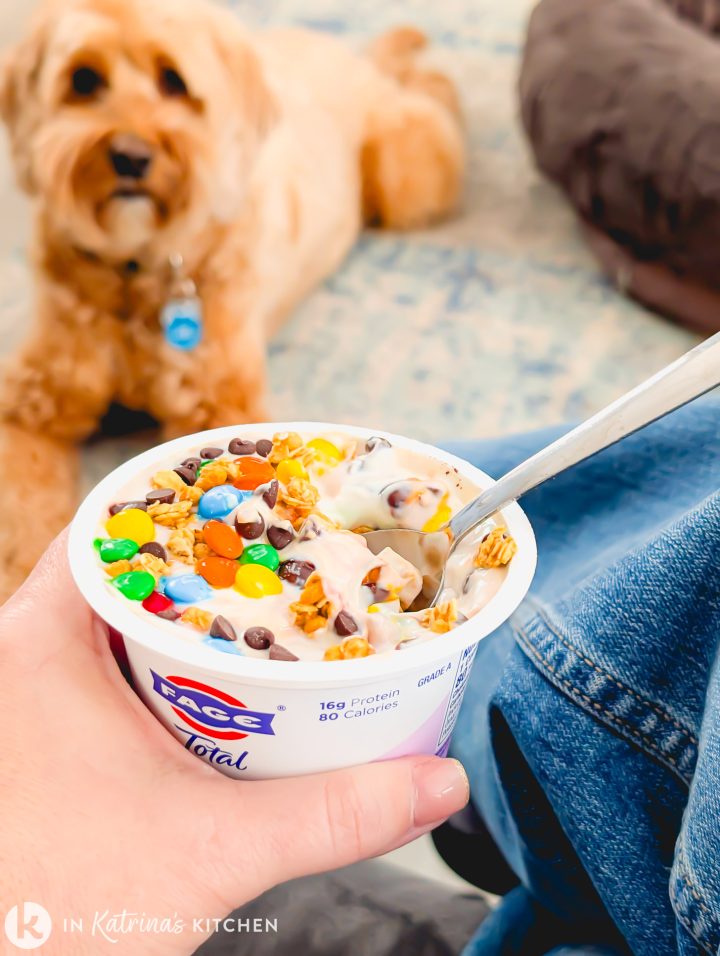 A person holding a single-serve container of FAGE Total Greek yogurt, topped with a mix of high-protein Monster Cookie Dough dip ingredients including colorful M&Ms, chocolate chips, and peanut butter cookie dough, with a labradoodle puppy in the background.