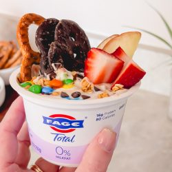 A hand holding a single-serve tub of FAGE Total 0% Greek yogurt topped with monster cookie dough dip ingredients, including M&Ms, chocolate chips, pretzel twists, sliced strawberries, and a chocolate cookie.